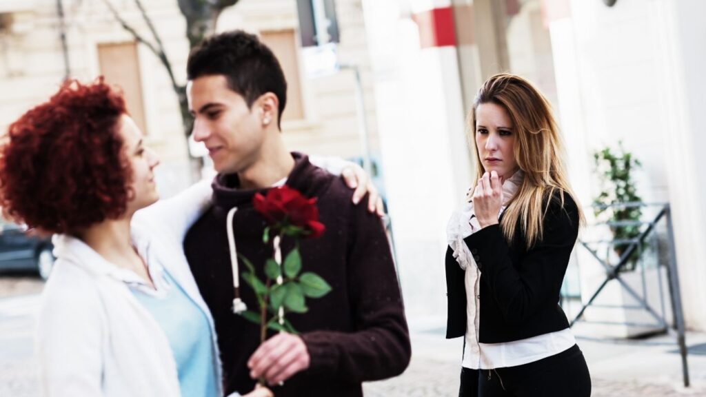 afastamento de rivais como funciona: casal abraçados enquanto homem segura uma rosa vermelha e uma outra mulher atrás olhando para eles.